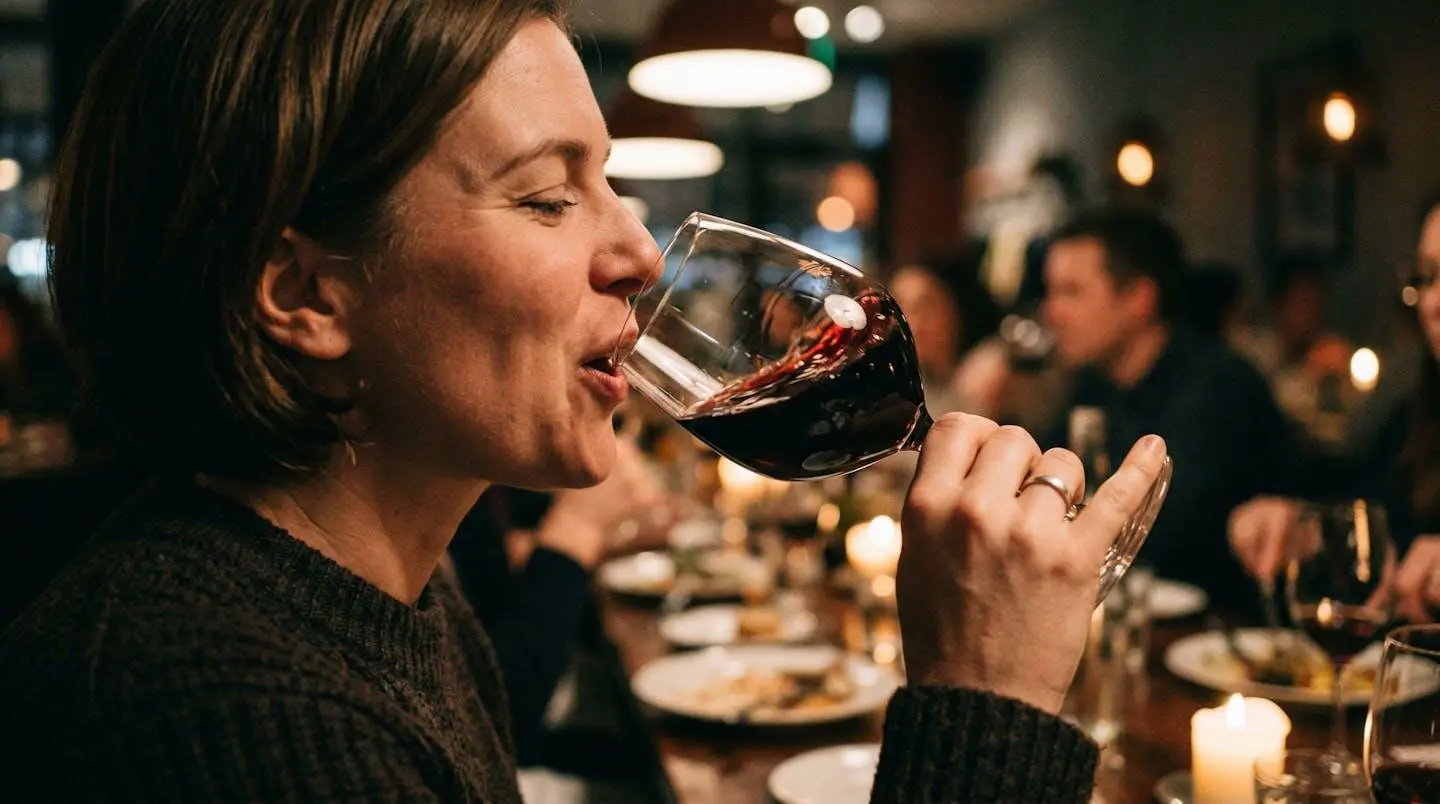 Une personne de profil incline doucement un verre de vin rouge vers ses lèvres, avec le mouvement du liquide visible et un arrière-plan flouté de table dressée